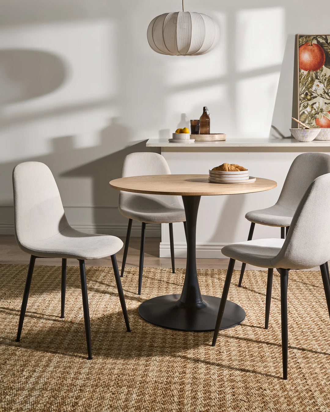 Light beige dining chairs arranged around a round wood-top pedestal table in a bright, modern dining room with warm sunlight, a woven rug, minimalist décor, and soft neutral accents.