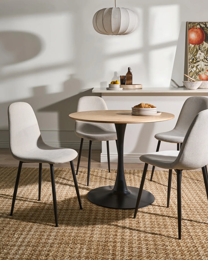 Light beige dining chairs arranged around a round wood-top pedestal table in a bright, modern dining room with warm sunlight, a woven rug, minimalist décor, and soft neutral accents.