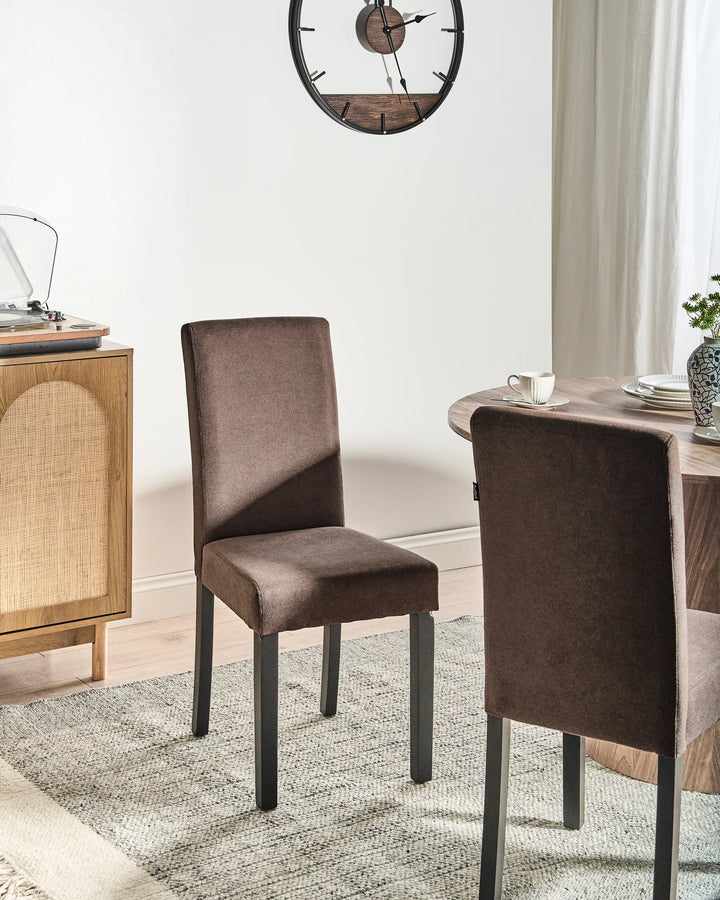 Brown dining chairs styled around a round wooden dining table on a textured rug, with a rattan-front cabinet, wall clock, and soft neutral décor creating a warm, contemporary setting.