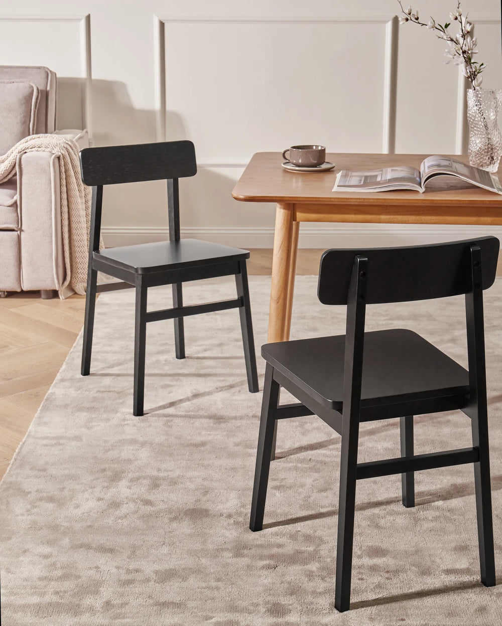 Black wooden dining chairs styled around a light wood dining table in a warm, modern dining space with neutral décor, soft lighting, a textured rug, and simple tabletop accents.