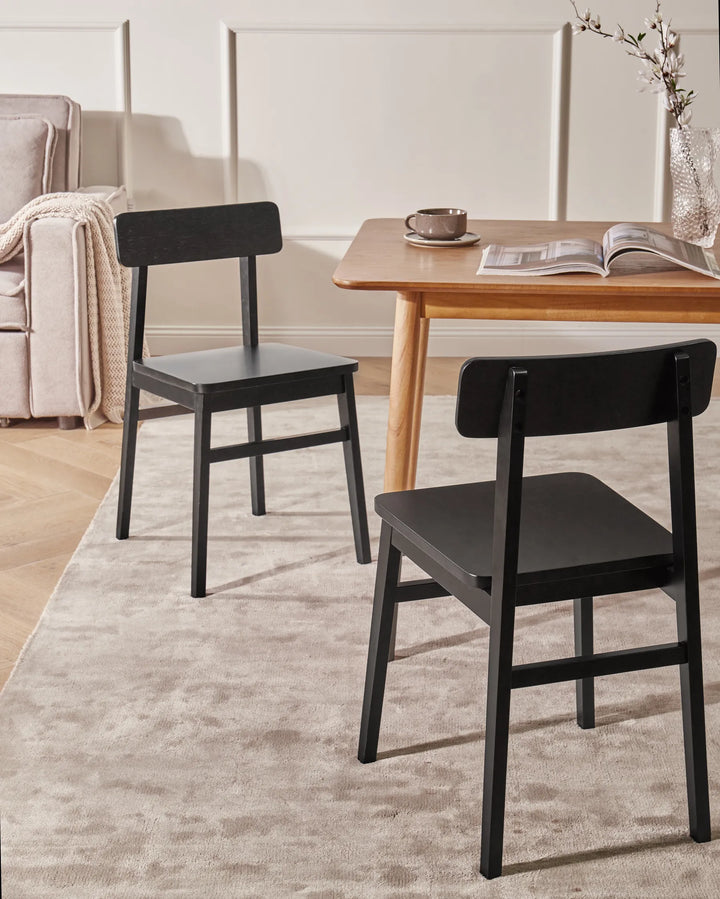 Black wooden dining chairs styled around a light wood dining table in a warm, modern dining space with neutral décor, soft lighting, a textured rug, and simple tabletop accents.