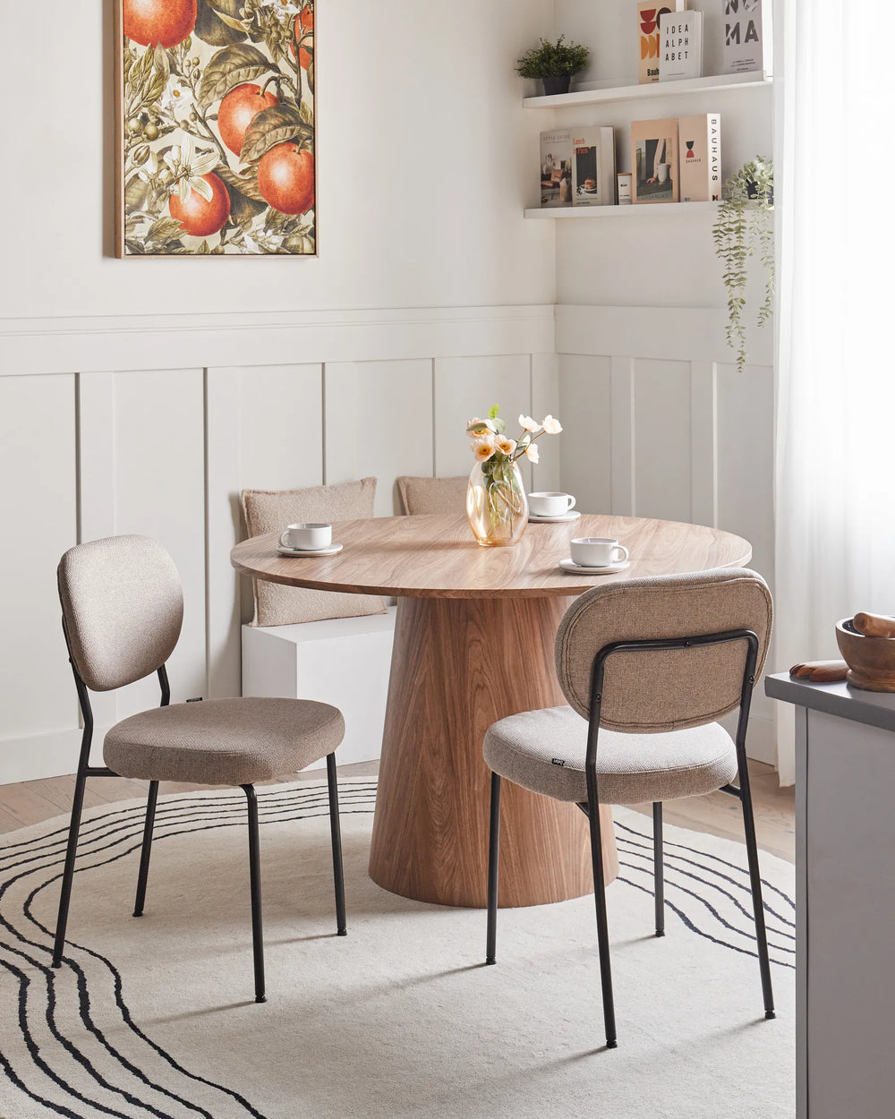 Beige fabric dining chairs paired with a round wooden pedestal table in a bright, modern dining room featuring neutral décor, wall shelving, floral accents, and warm natural light.