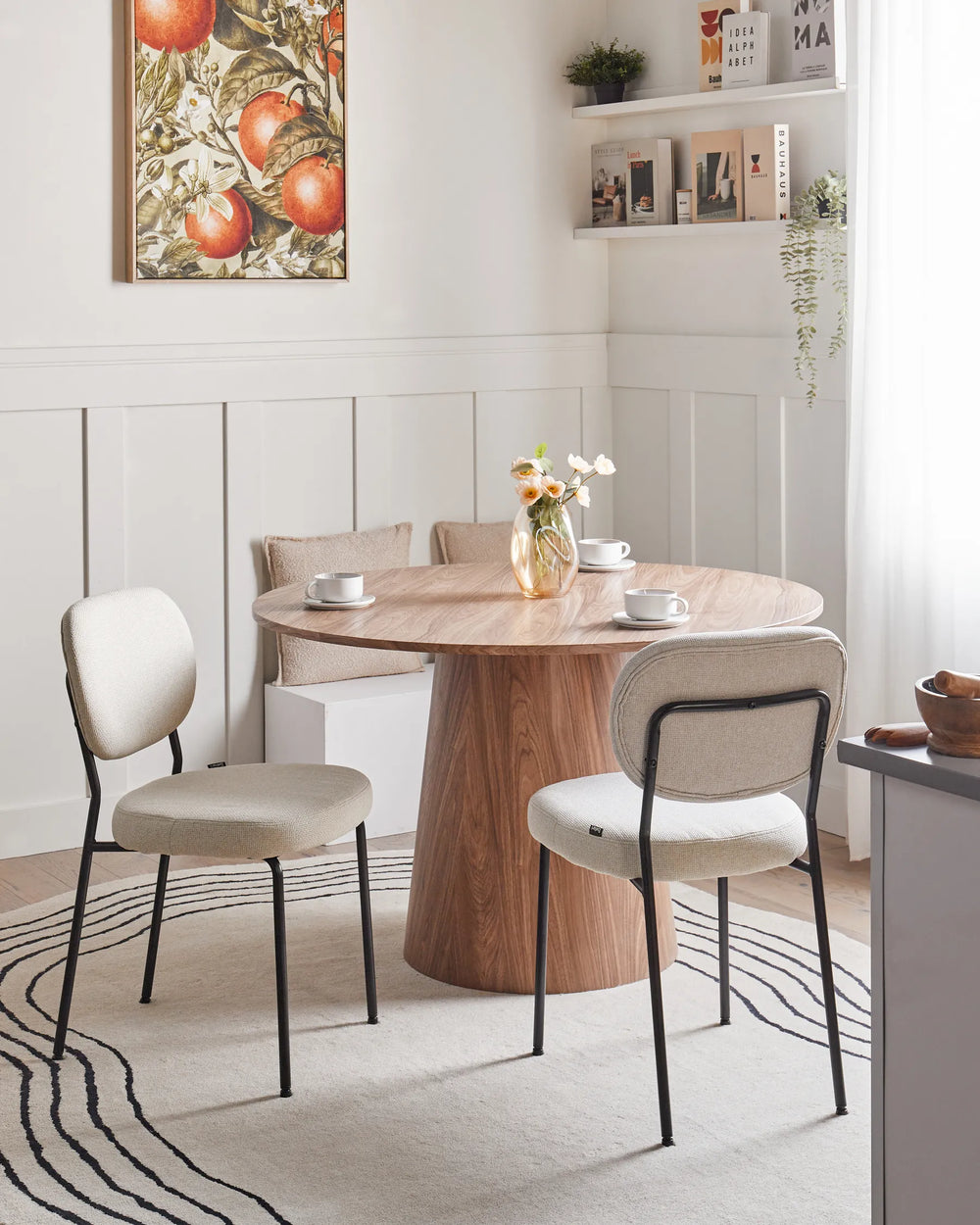 Beige upholstered dining chairs styled around a round wooden pedestal table in a bright, modern dining space featuring soft neutral décor, wall shelving, floral accents, and natural light.