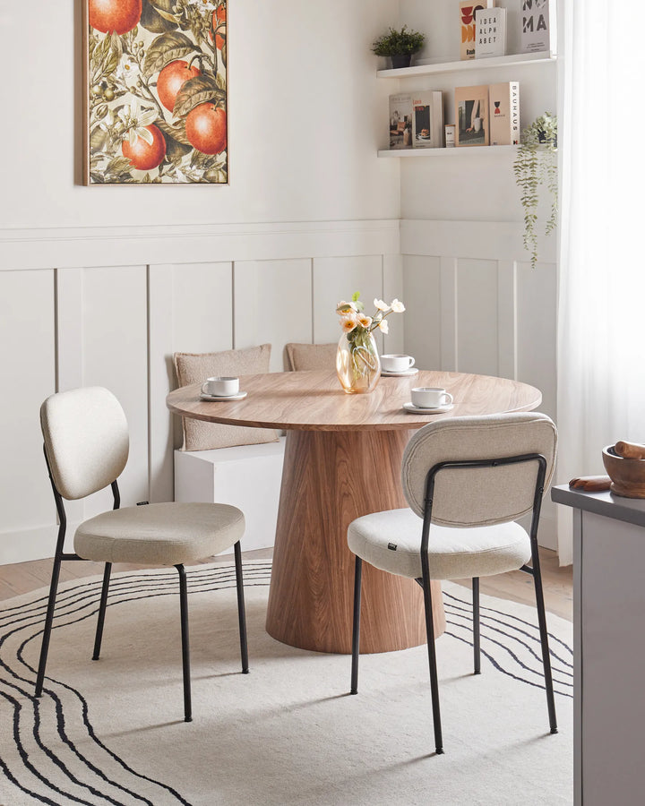 Beige upholstered dining chairs styled around a round wooden pedestal table in a bright, modern dining space featuring soft neutral décor, wall shelving, floral accents, and natural light.