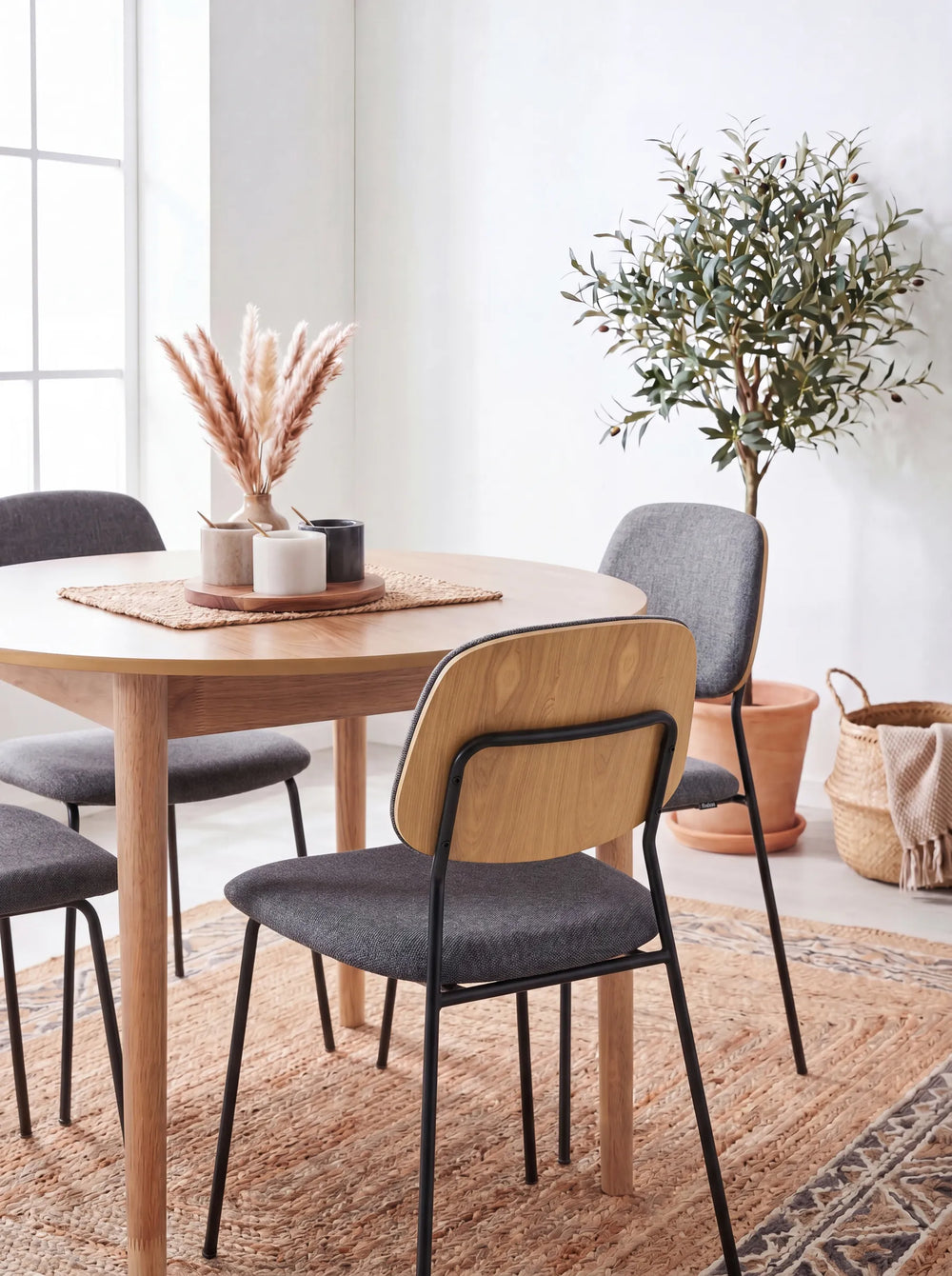 Dark grey dining chairs around a round wooden table with a woven rug, decorative vase, olive tree, and bright natural light.