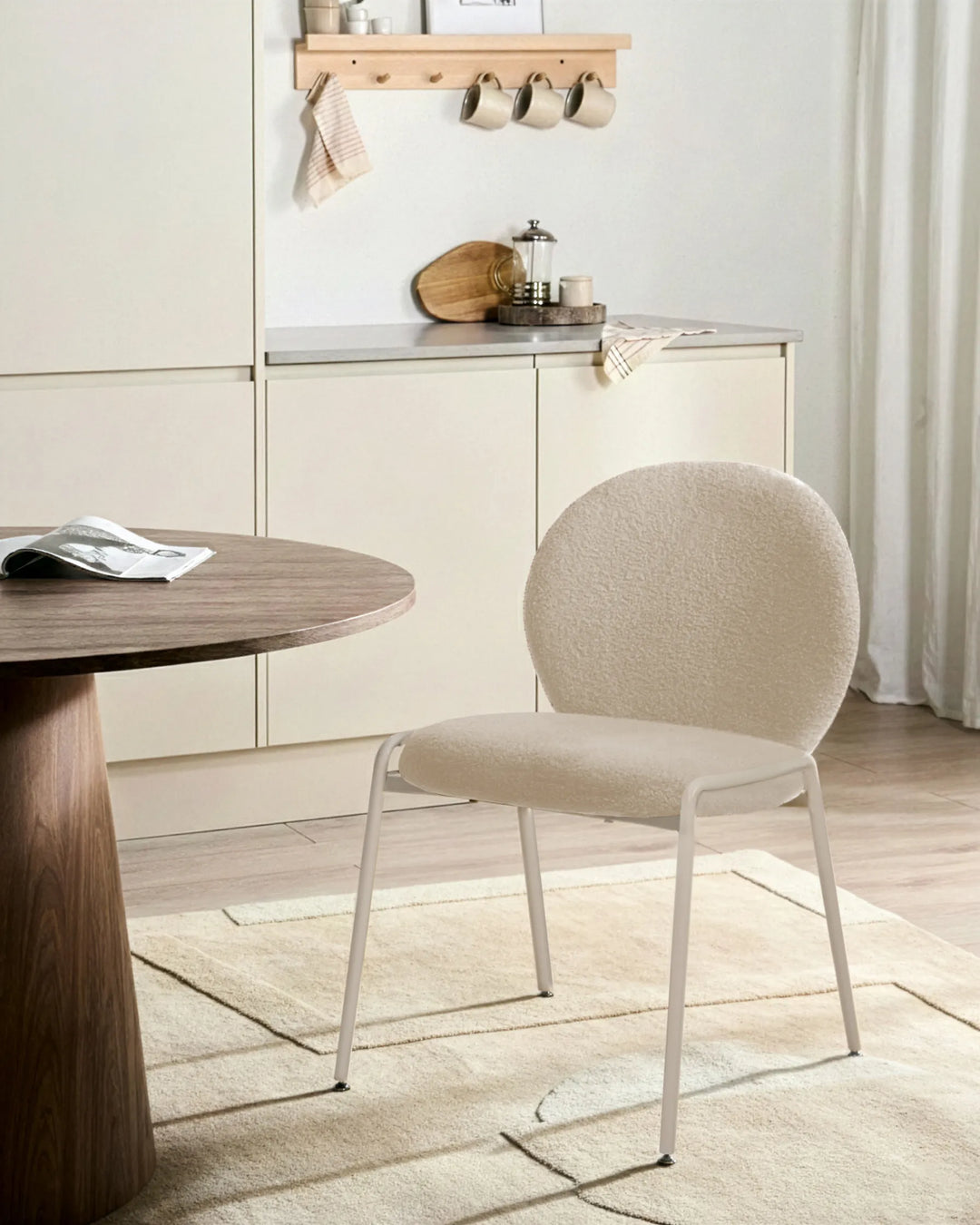 A pair of light beige boucle chairs at a modern dining table in a bright, minimalist room with warm wooden accents and neutral decor.