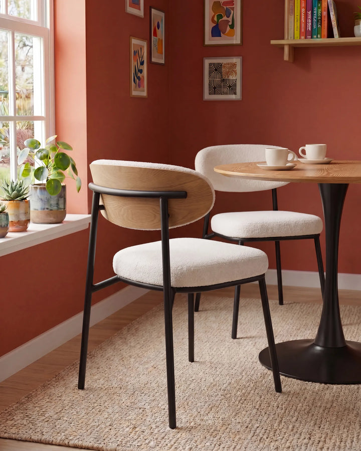 Cream bouclé dining chairs around a round wooden dining table in a warm dining room with terracotta walls, framed artwork, indoor plants, and soft natural light.