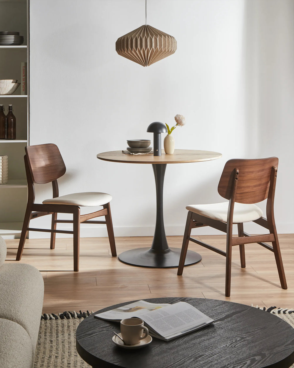 Walnut dining chairs with cream seats around a round dining table in a modern dining space with a black pedestal base, wooden pendant light, and minimalist décor.