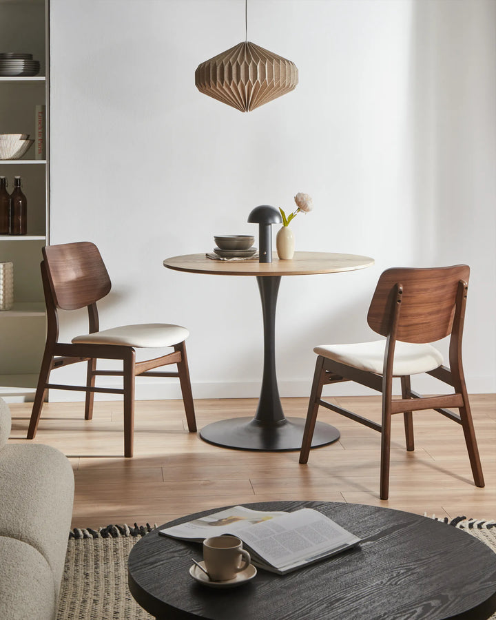Walnut dining chairs with cream seats around a round dining table in a modern dining space with a black pedestal base, wooden pendant light, and minimalist décor.
