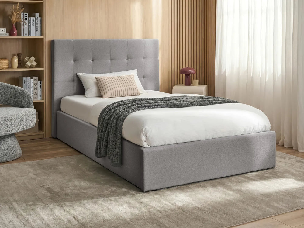 Grey upholstered bed in a room with a wooden floor and a bookshelf.