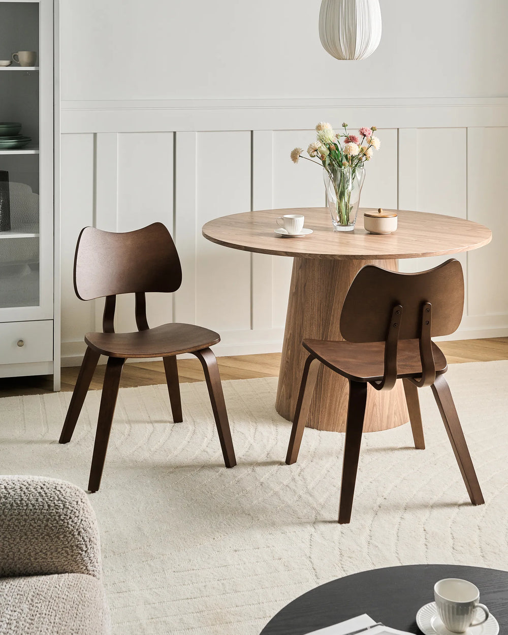 Dark walnut dining chairs styled around a round wooden dining table in a warm minimalist dining space featuring soft neutral tones, floral accents, natural textures, and cozy contemporary styling.