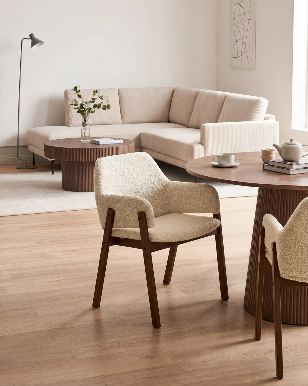 Cream bouclé armchairs with walnut frames around a round wooden table in a bright, modern living space featuring a neutral sofa, coffee tables, and minimalist décor.