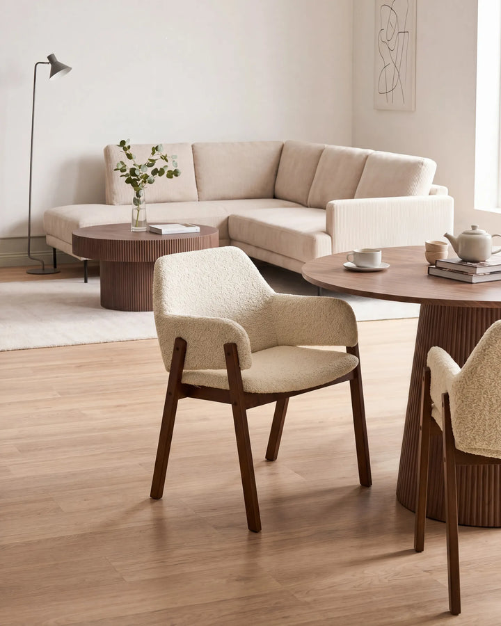 Cream bouclé armchairs with walnut frames around a round wooden table in a bright, modern living space featuring a neutral sofa, coffee tables, and minimalist décor.