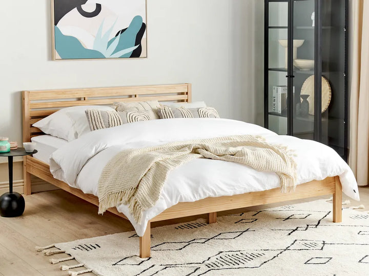 Light brown pine wood bed frame in a simple bedroom setting with a neutral wall, soft bedding, and minimal decor in the background.