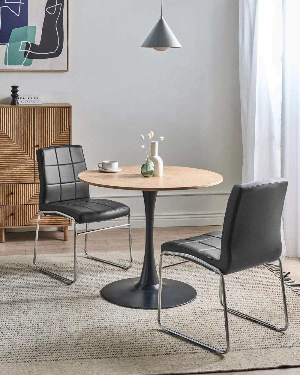 Black cantilever dining chairs paired with a round oak table and black pedestal base, styled in a bright contemporary dining space with a textured wood sideboard, neutral rug, minimalist pendant light, and soft natural daylight.