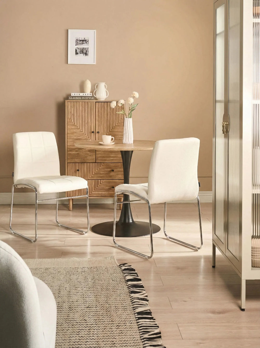 White dining chairs styled around a round wood-top table with a black pedestal base, set in a warm neutral interior featuring a textured wood sideboard, light wood flooring, and soft beige walls.