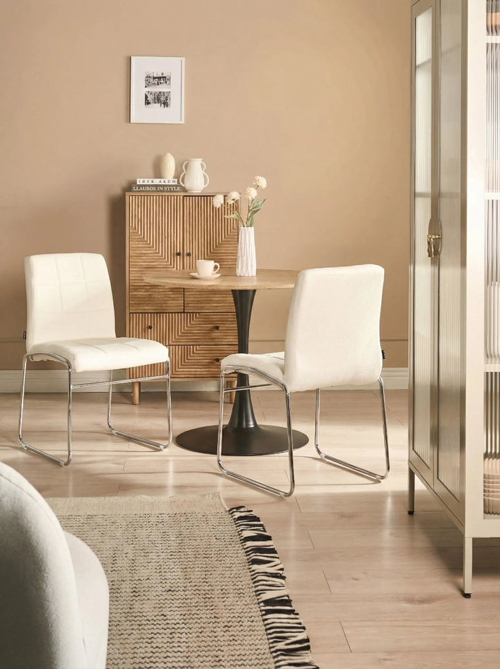 White dining chairs styled around a round wood-top table with a black pedestal base, set in a warm neutral interior featuring a textured wood sideboard, light wood flooring, and soft beige walls.