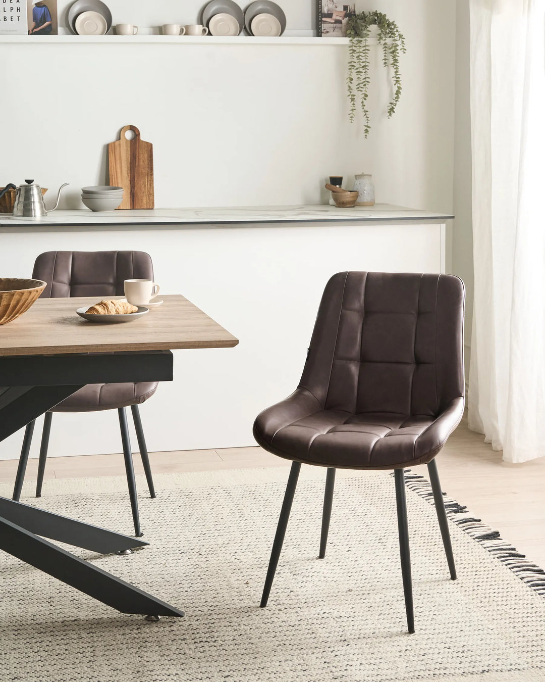 Brown faux leather dining chairs styled around a wooden dining table in a bright modern dining space with open shelving, hanging greenery, light flooring, and a textured neutral rug.
