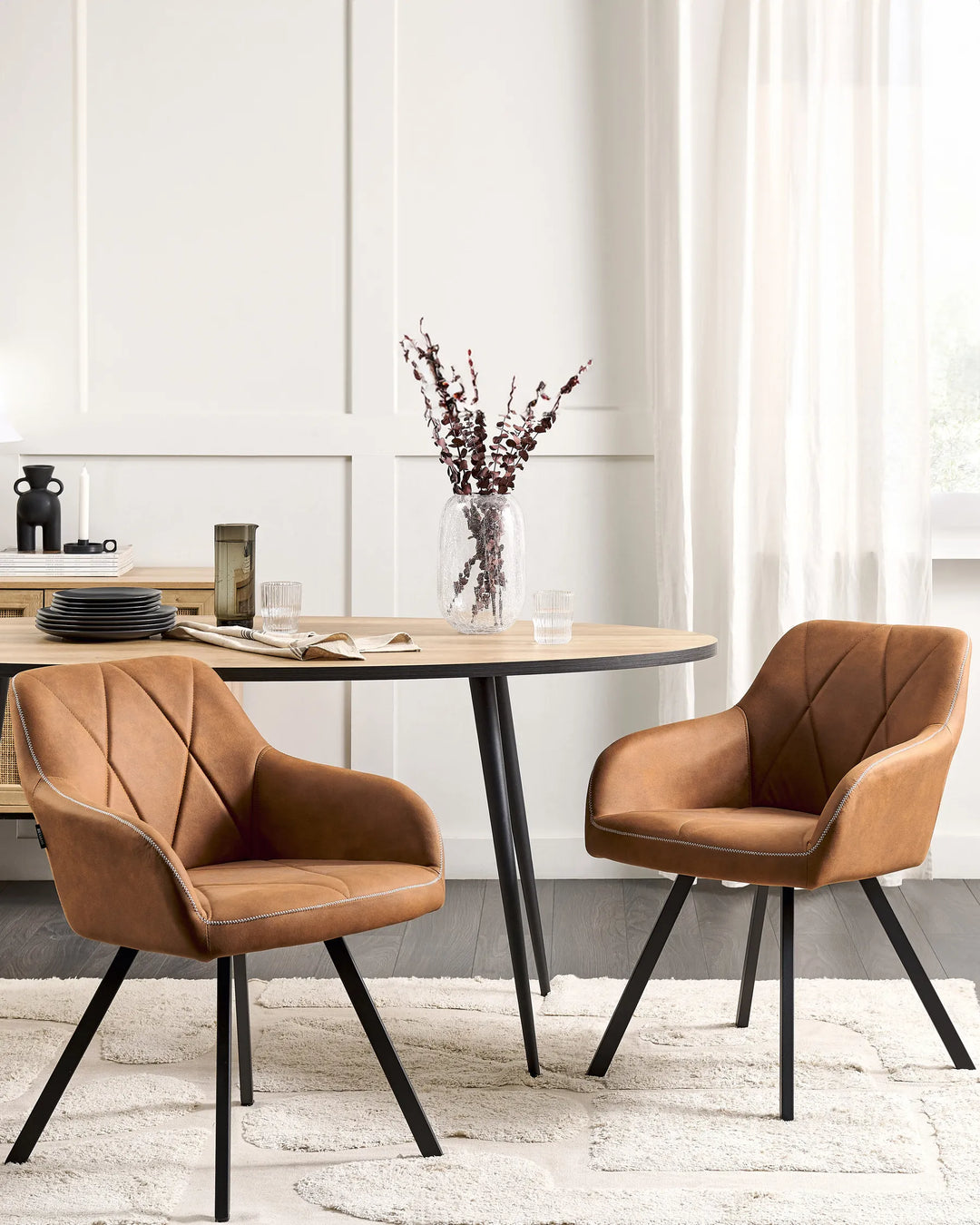 Stylish dining space with matching cognac-brown armchairs around a light wood table, complemented by black metal accents, soft neutral textiles, and elegant decorative accessories.