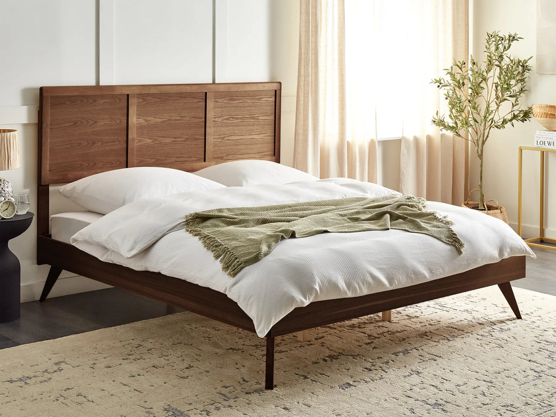 A dark ash wood bed with a solid headboard in a cozy bedroom, featuring neutral decor and a leafy plant against warm-toned walls and floor.