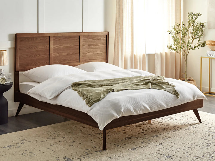 A dark ash wood bed with a solid headboard in a cozy bedroom, featuring neutral decor and a leafy plant against warm-toned walls and floor.