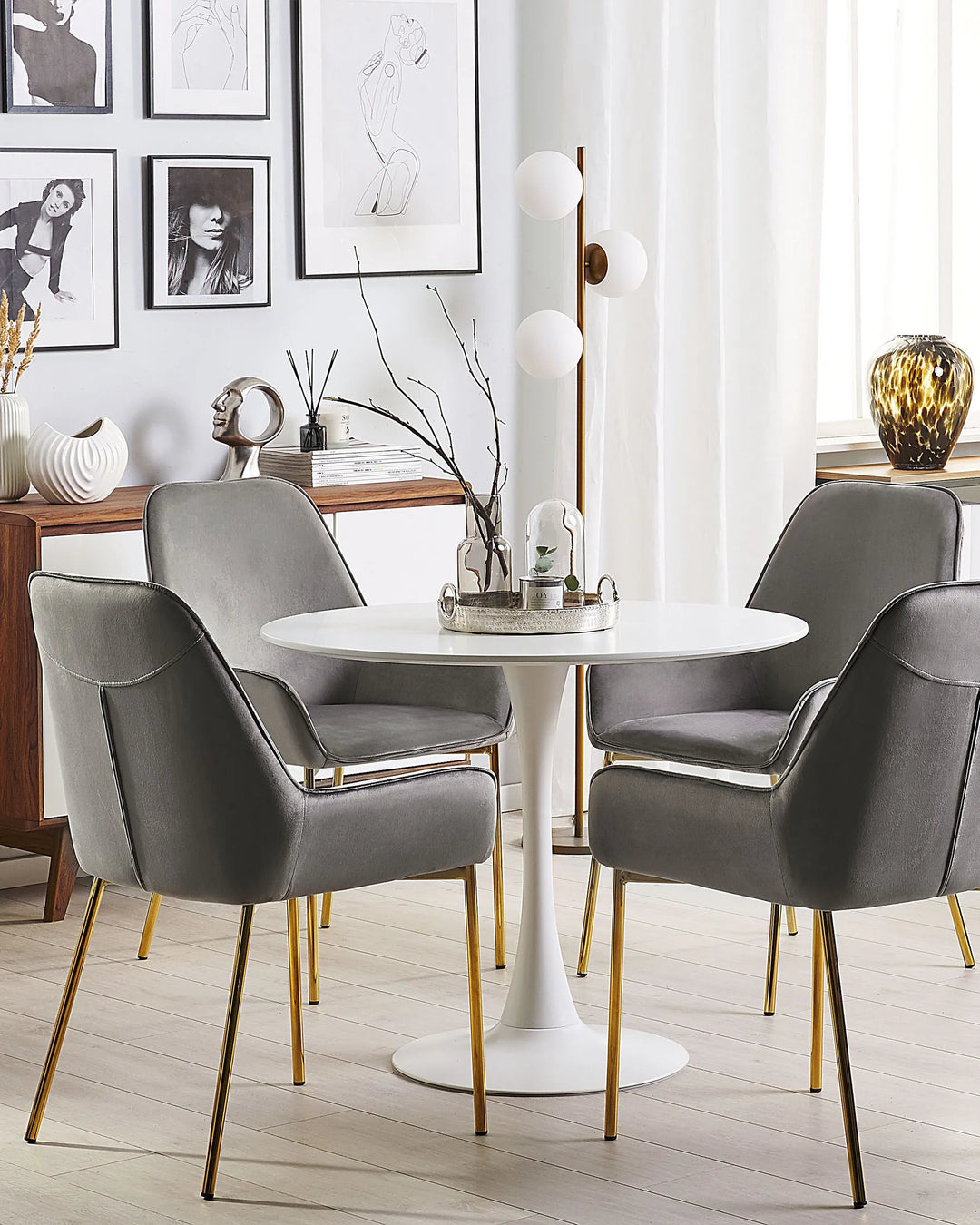 Stylish dining area featuring grey velvet dining chairs with gold accents around a round white pedestal table, complemented by modern artwork, warm metallic décor, and bright natural light for a refined, upscale aesthetic.