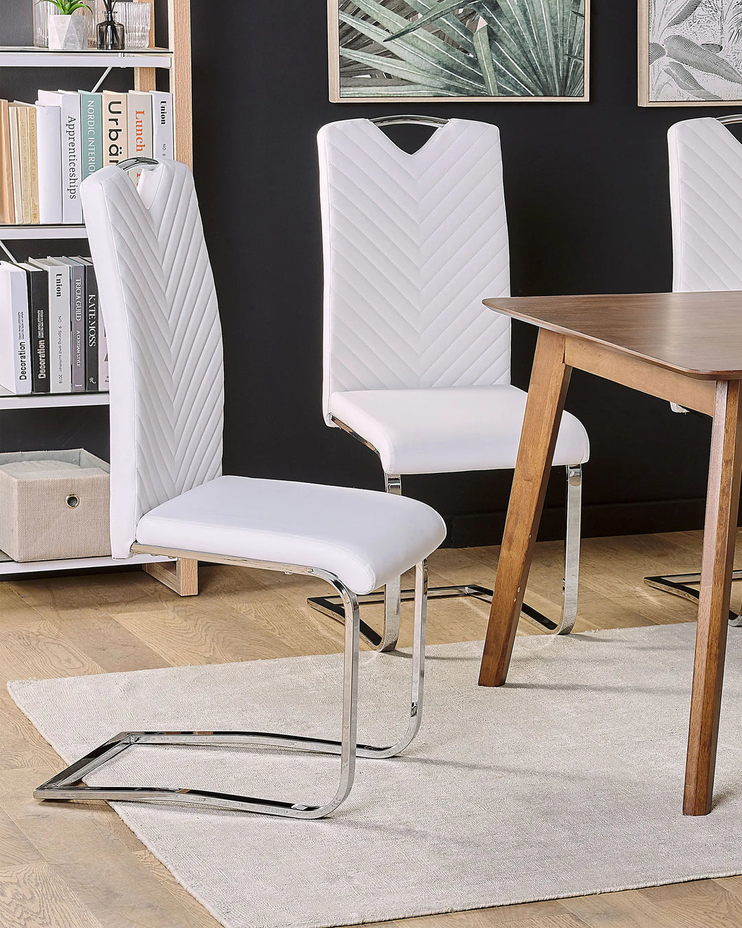 White faux leather cantilever dining chairs styled around a wooden dining table in a contemporary dining room with a black accent wall, botanical wall art, open shelving, light wood flooring, and a soft neutral area rug.