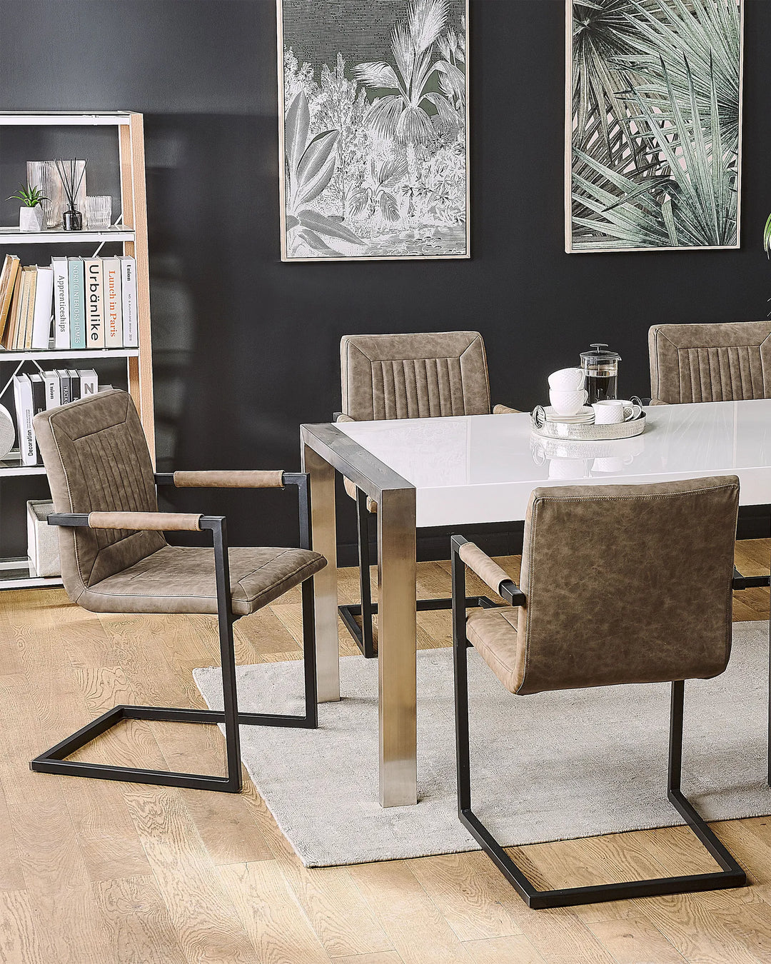 Brown dining chairs arranged around a glossy white dining table on a light rug, set against dark accent walls with botanical artwork, open shelving, and modern décor.