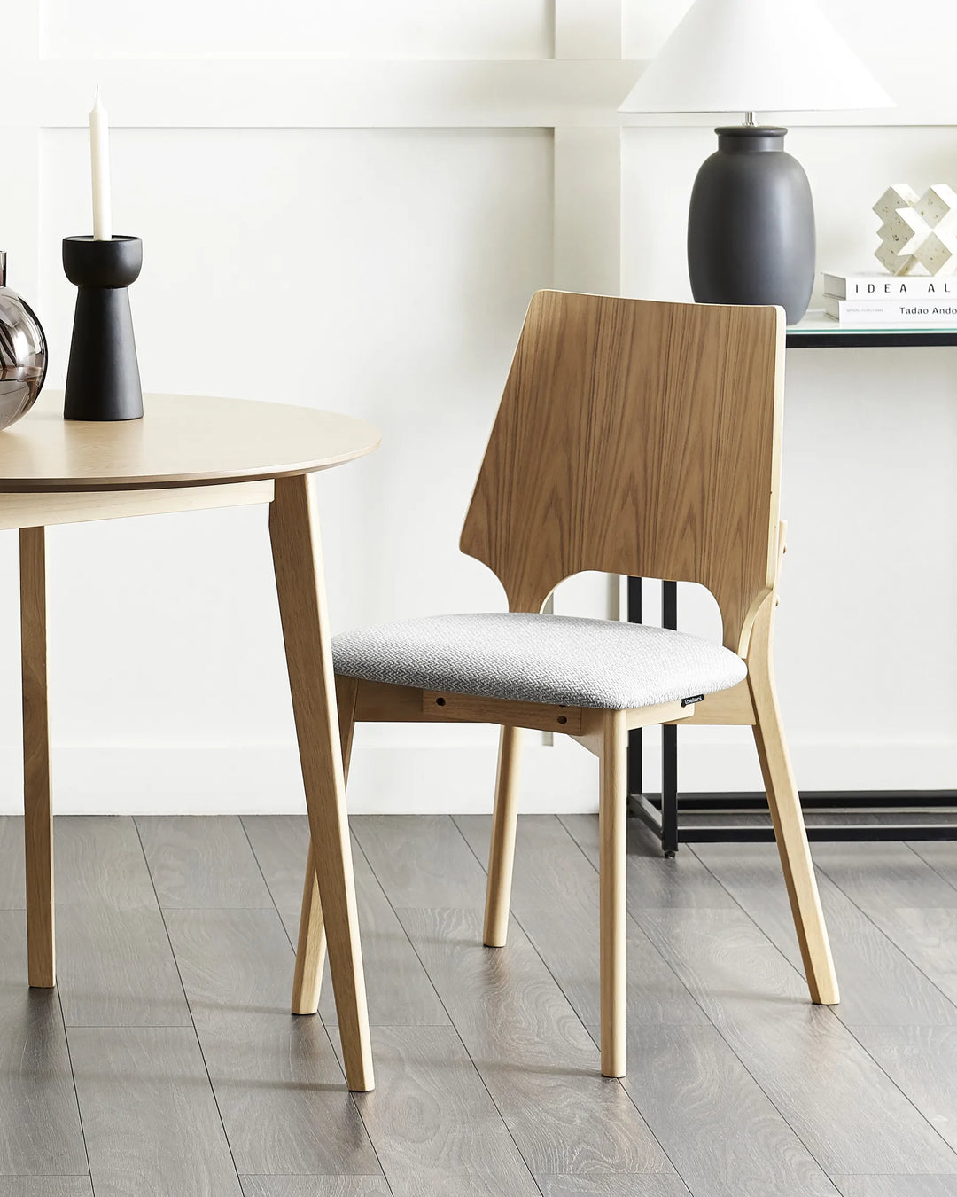Styled within a bright, minimalist dining setting, this upholstered dining chair brings warmth and character through its contrasting wood tones and clean architectural lines. The soft grey seat and natural finish create a welcoming atmosphere that blends seamlessly with modern, Scandinavian, and contemporary interiors.