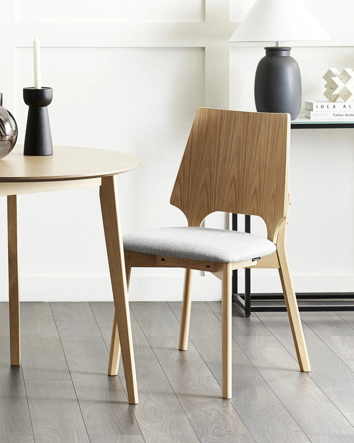 Styled within a bright, minimalist dining setting, this upholstered dining chair brings warmth and character through its contrasting wood tones and clean architectural lines. The soft grey seat and natural finish create a welcoming atmosphere that blends seamlessly with modern, Scandinavian, and contemporary interiors.