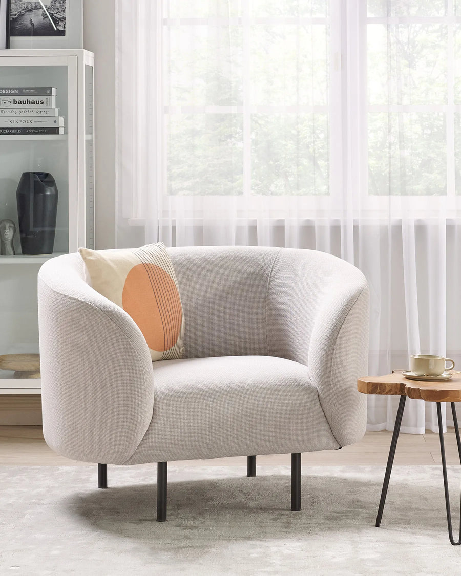 Modern beige armchair with a decorative pillow in a bright room with white curtains.
