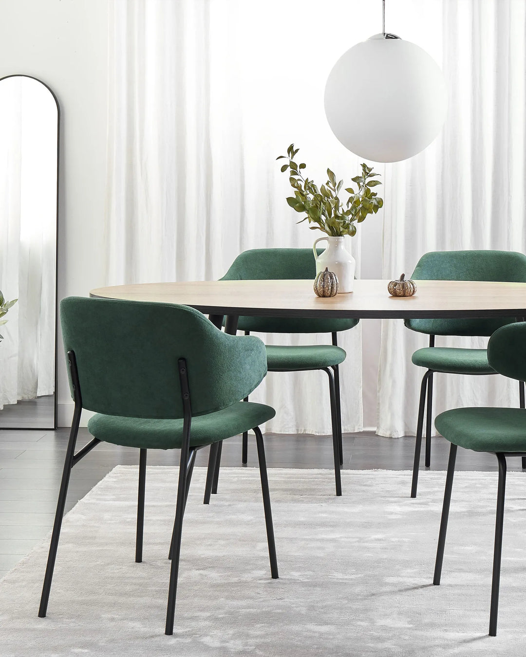 Green upholstered dining chairs arranged around a light wood dining table in a bright modern dining room with sheer white curtains, a round pendant light, decorative greenery, a floor mirror, and a soft neutral rug.