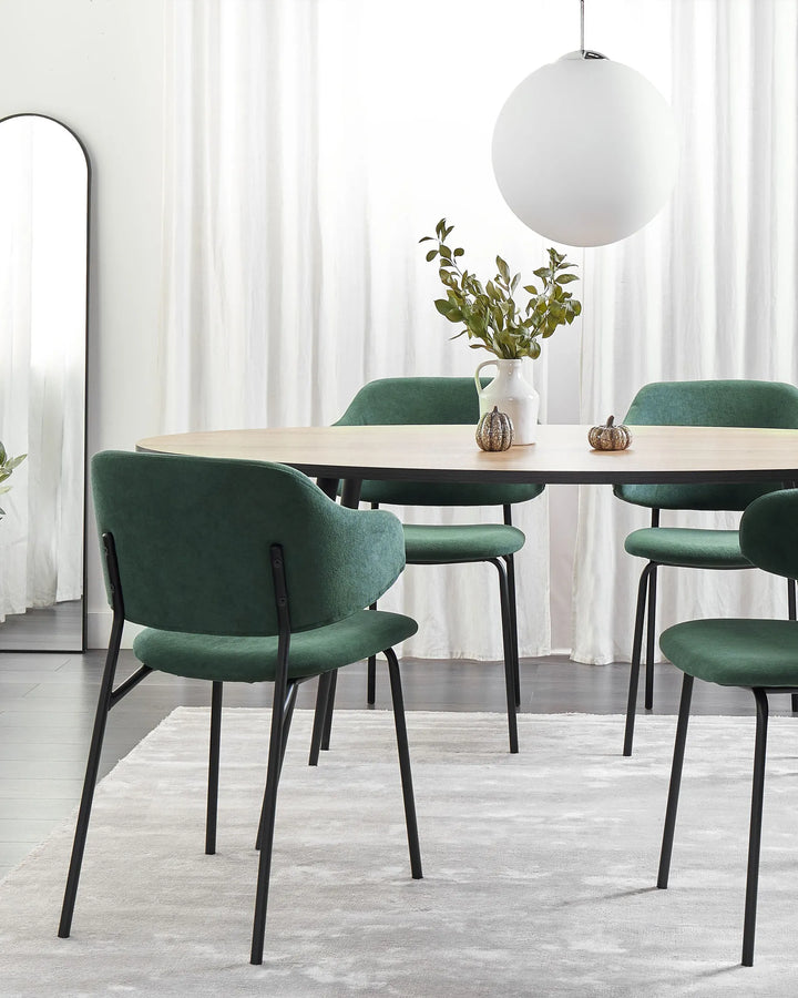 Green upholstered dining chairs arranged around a light wood dining table in a bright modern dining room with sheer white curtains, a round pendant light, decorative greenery, a floor mirror, and a soft neutral rug.