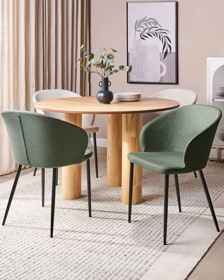 Green upholstered dining chairs styled around a round light wood dining table with a black vase, stacked plates, framed botanical artwork, neutral curtains, and a textured cream rug.