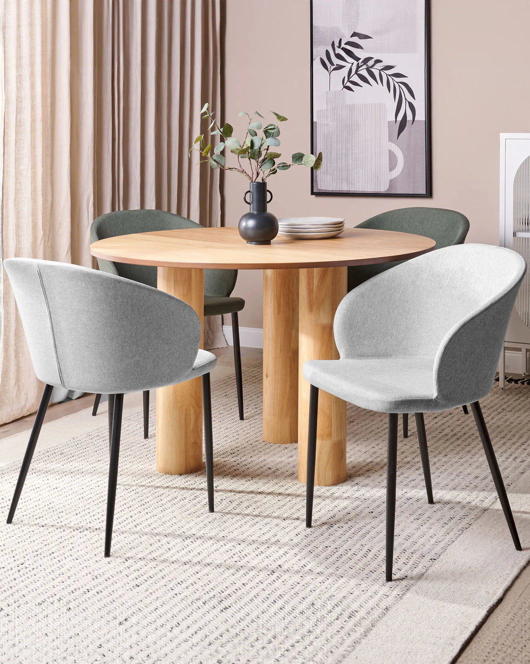 Light grey upholstered dining chairs paired with a round light wood dining table, black vase with eucalyptus stems, stacked plates, framed botanical wall art, beige curtains, and a textured cream rug in a modern dining room.