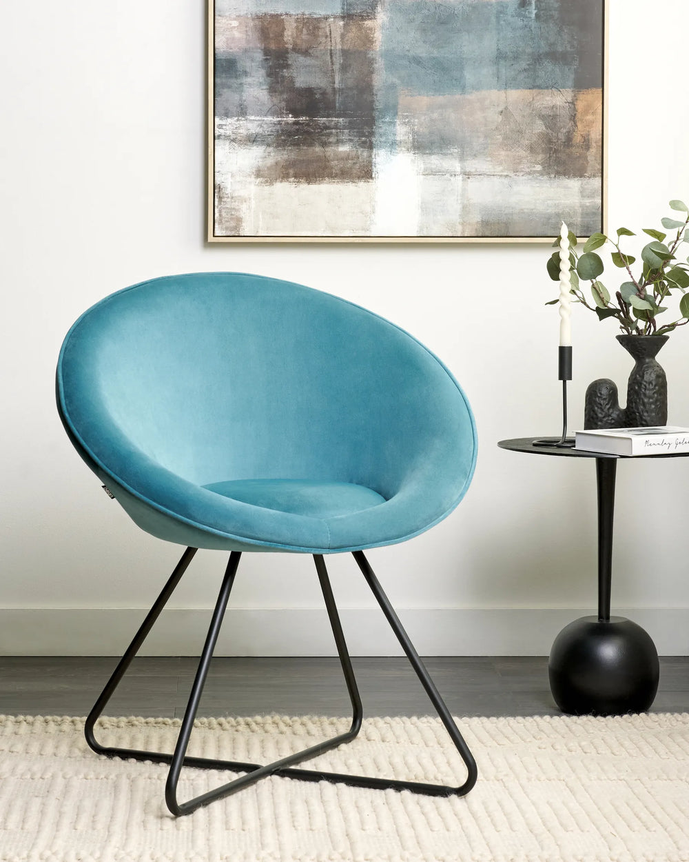 Blue velvet accent chair styled beside a black side table with candles, greenery, abstract wall art, and a textured cream rug.