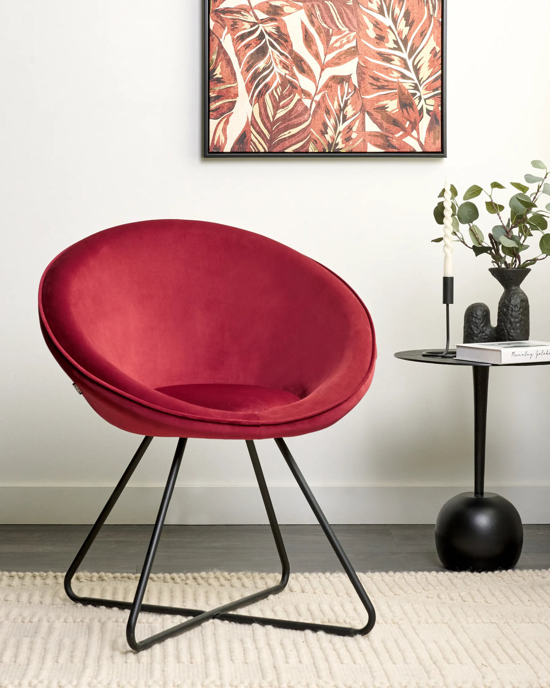 Red velvet accent chair styled beside a black side table with candles, greenery, framed botanical artwork, and a textured cream rug.