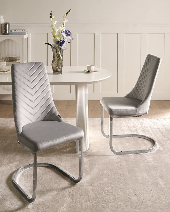 Light grey velvet cantilever dining chairs paired with a round white dining table in a bright, contemporary dining room with soft neutral décor and fresh flowers.