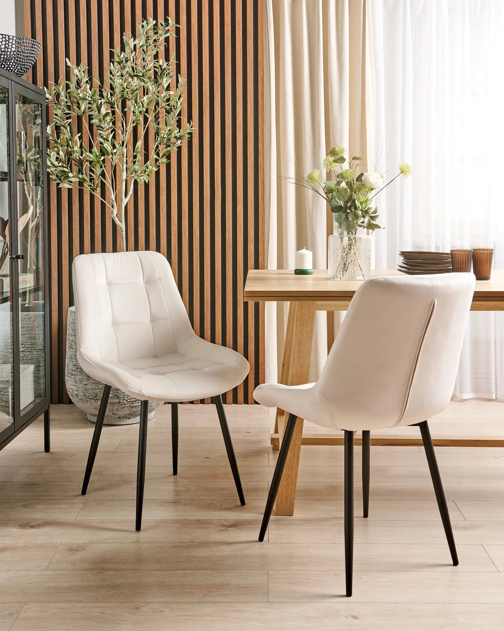 White faux leather dining chairs styled around a wooden dining table in a modern dining room with wood slat wall panels, decorative greenery, light flooring, and soft natural light.