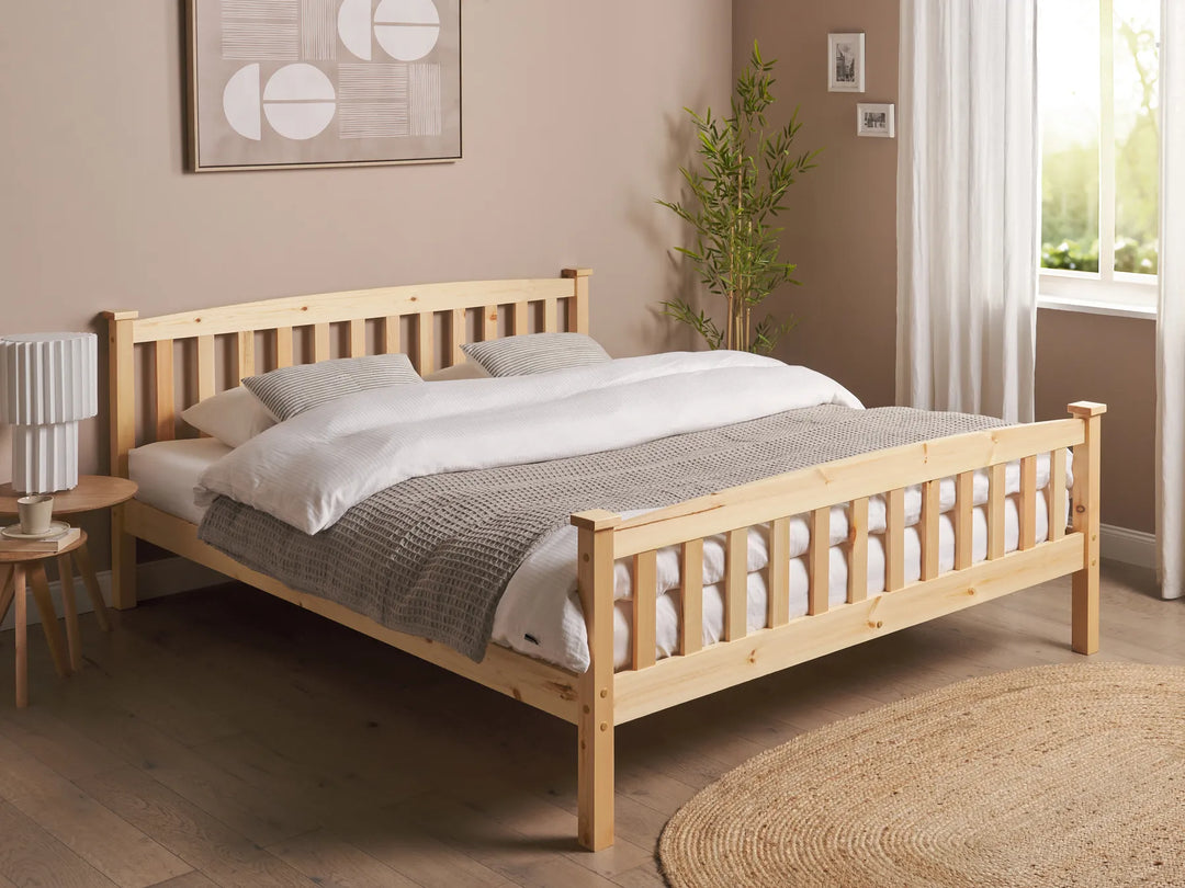 Wooden bed with white bedding in a room with a lamp, plant, and artwork on the wall.