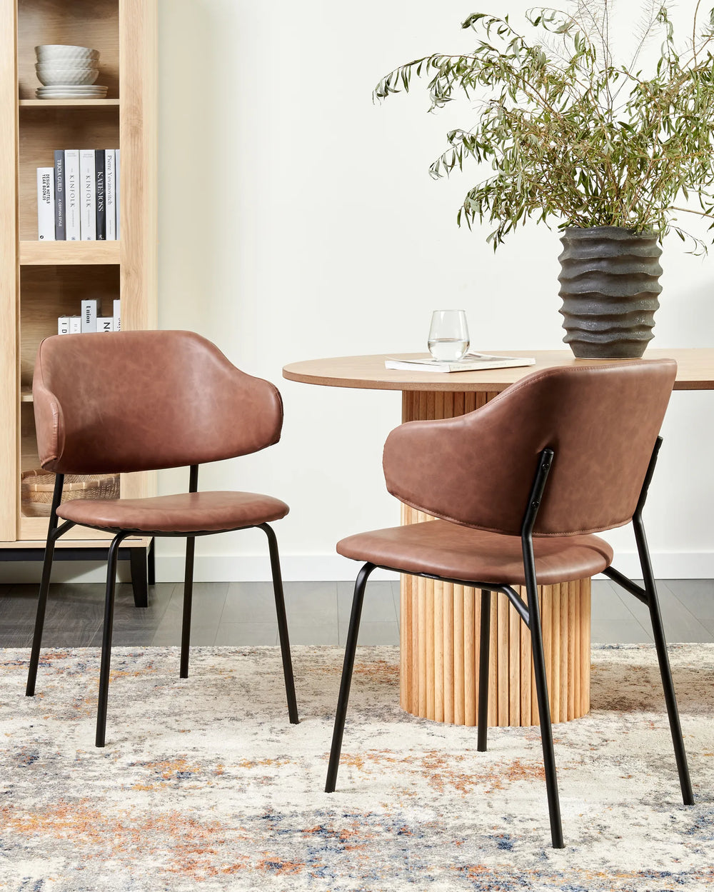 Brown faux leather dining chairs styled around a round fluted wood dining table in a warm contemporary dining space with a light wood bookcase, decorative plant, textured area rug, and neutral-toned walls.