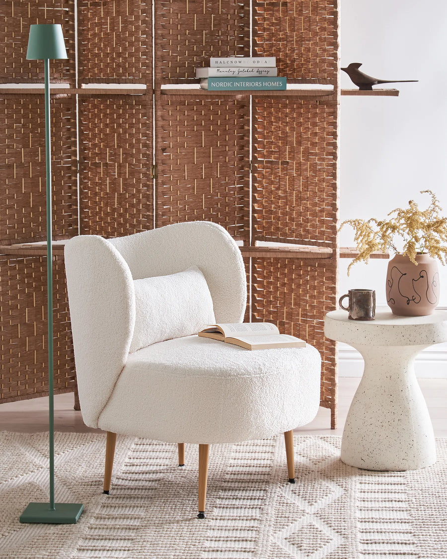 White armchair in a room with a woven wall, floor lamp, and small table with decor items.