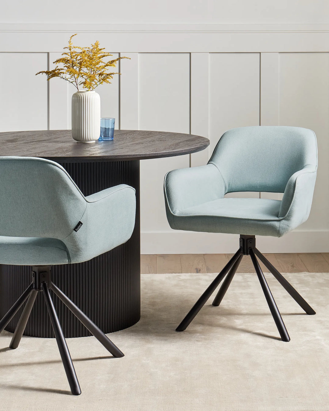 Light blue swivel dining chairs styled around a black round dining table with a vase of yellow flowers, blue glass, cream rug, and white panelled wall backdrop.