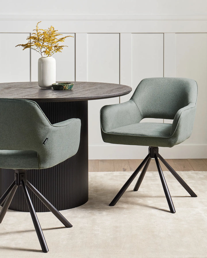 Green swivel dining chairs styled around a black round dining table with a white vase of yellow flowers, decorative bowl, cream rug, and white panelled wall backdrop.