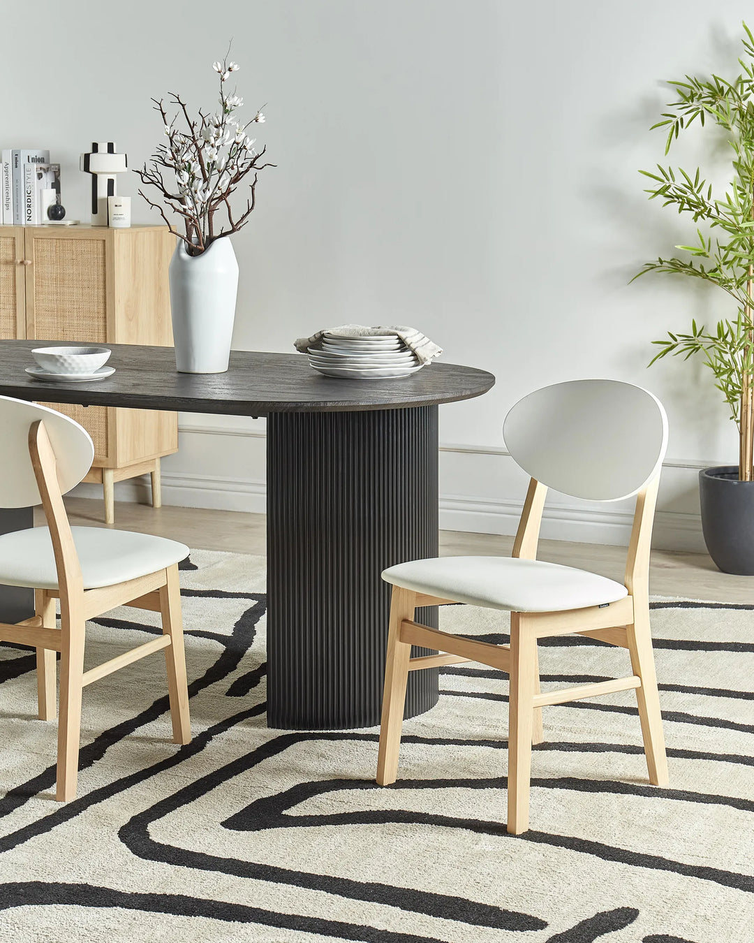 Light wood dining chairs with soft grey upholstery styled around a dark round dining table in a contemporary dining space featuring a patterned rug, neutral décor, indoor greenery, and minimalist Scandinavian-inspired styling.
