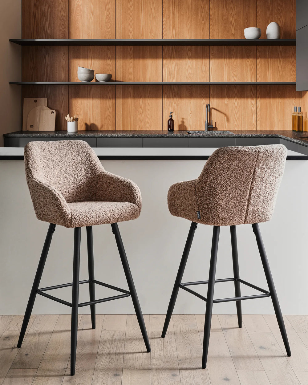 Taupe bouclé bar chairs with rounded backrests and slanted black metal legs around a bar counter in a contemporary kitchen or dining space with modern décor.