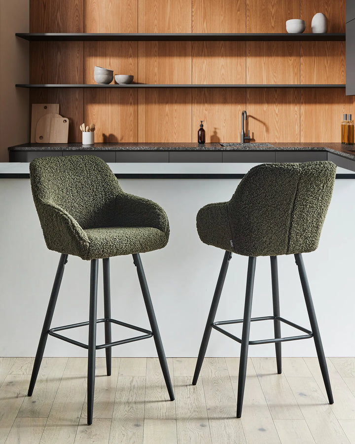 Dark green bouclé bar chairs with rounded backrests and slanted black metal legs around a bar counter in a contemporary kitchen or dining space with modern décor.