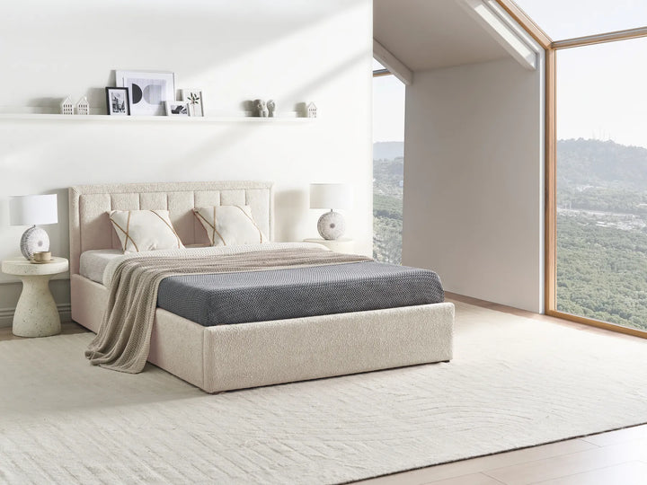 Beige upholstered bed with gray bedding in a bright bedroom with large windows.