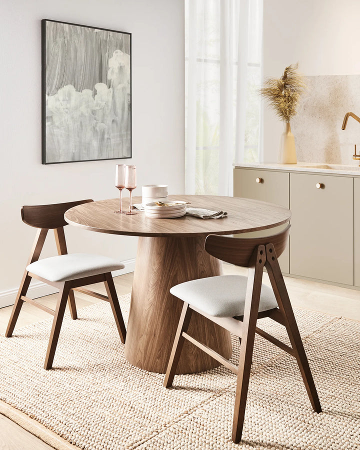 Placed around a warm wood pedestal dining table, this modern chair adds richness and contrast through its dark wood finish and soft neutral upholstery. The clean Scandinavian-inspired silhouette complements contemporary, mid-century, and minimalist interiors, making it an elegant choice for dining spaces, breakfast nooks, and open-plan living areas.