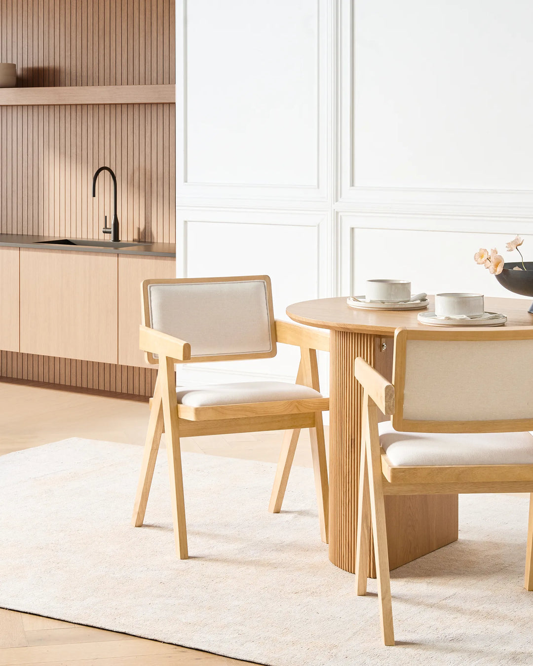 Showcased in a bright, sophisticated dining setting, this upholstered wooden armchair adds warmth, comfort, and understated elegance to the room. The natural oak frame pairs beautifully with the neutral fabric upholstery, creating a calm and inviting atmosphere. Positioned around a coordinating round dining table, the chair complements contemporary interiors while offering a comfortable seating experience for everyday dining and entertaining.
